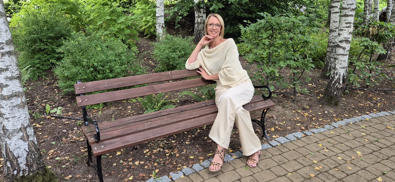 Nathalie assise sur un banc dans un sous-bois de bouleaux, tenue crème